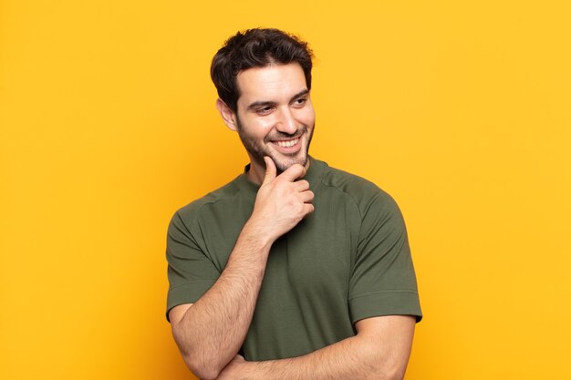 Hombre guapo joven sonriendo con una expresión feliz y segura con la mano en la barbilla, preguntándose y mirando hacia un lado