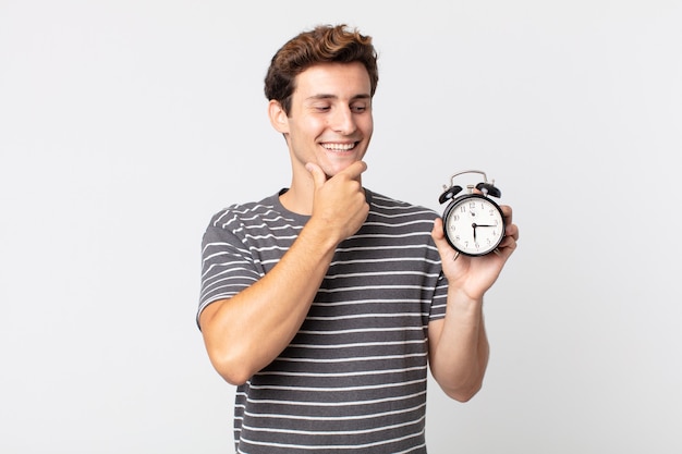 Hombre guapo joven sonriendo con una expresión feliz y segura con la mano en la barbilla y sosteniendo un reloj despertador