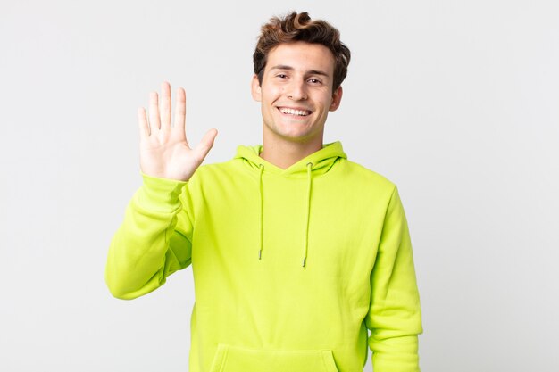 Hombre guapo joven sonriendo feliz y alegremente, saludando con la mano, dándote la bienvenida y saludándote, o diciéndote adiós