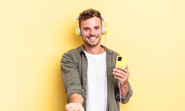 Hombre guapo joven sonriendo felizmente con amigable y ofreciendo y mostrando un concepto de concepto de auriculares y teléfono inteligente