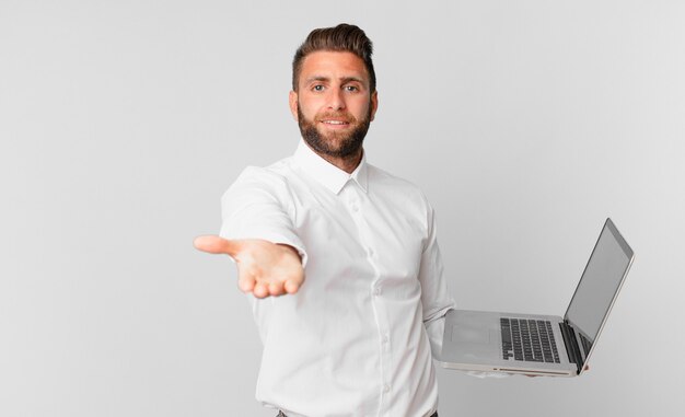 Hombre guapo joven sonriendo felizmente con amigable y ofreciendo y mostrando un concepto y sosteniendo una computadora portátil