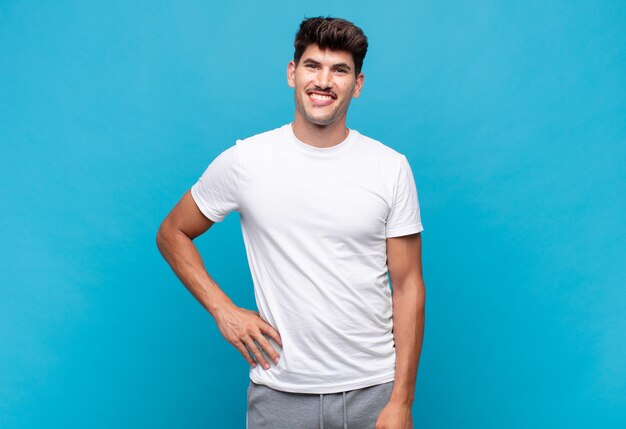 Foto hombre guapo joven sonriendo felizmente con una mano en la cadera y actitud confiada, positiva, orgullosa y amistosa