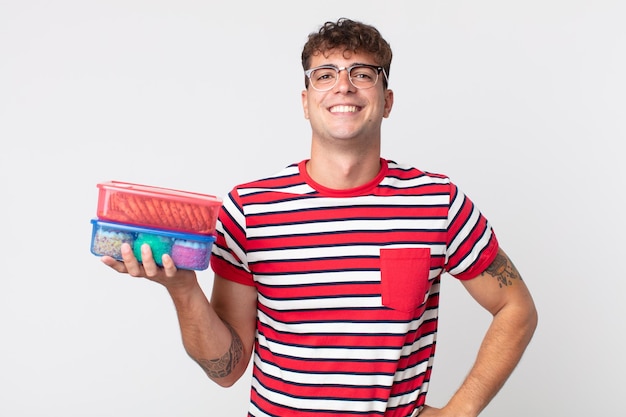 Hombre guapo joven sonriendo felizmente con una mano en la cadera y confiado y sosteniendo una lonchera