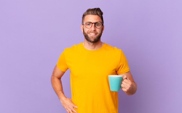 Hombre guapo joven sonriendo felizmente con una mano en la cadera y confiado. y sosteniendo una taza de café