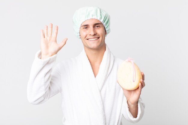 Hombre guapo joven sonriendo felizmente, saludando con la mano, dándote la bienvenida y saludándote con albornoz, gorro de ducha y una esponja