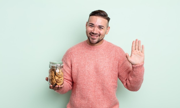 Hombre guapo joven sonriendo felizmente, saludando con la mano, dándote la bienvenida y saludándote. concepto de botella de galletas