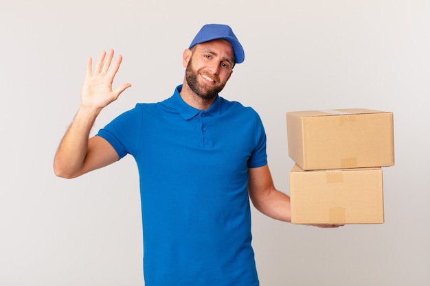 Hombre guapo joven sonriendo felizmente, saludando con la mano, dándote la bienvenida y saludándote. concepto de entrega de paquetes