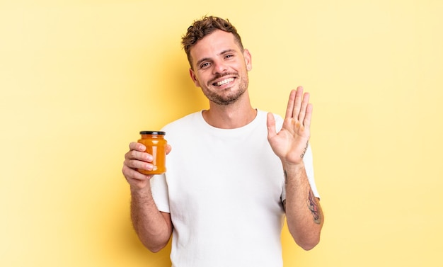 Hombre guapo joven sonriendo felizmente, saludando con la mano, dándote la bienvenida y saludándote. concepto de gelatina de durazno