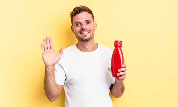 Hombre guapo joven sonriendo felizmente, saludando con la mano, dándote la bienvenida y saludándote. concepto de salsa de tomate