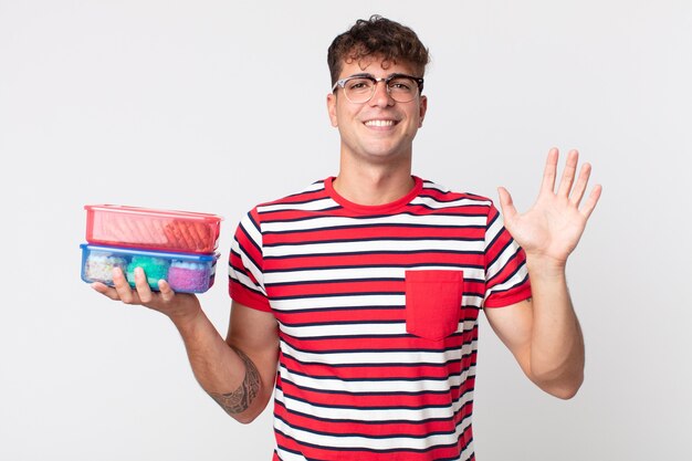 Hombre guapo joven sonriendo felizmente, saludando con la mano, dándote la bienvenida y saludándote y sosteniendo una lonchera