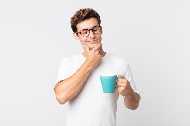 Hombre guapo joven sonriendo felizmente y soñando despierto o dudando y sosteniendo una taza de café