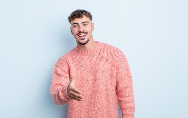 Hombre guapo joven sonriendo, luciendo feliz, confiado y amigable, ofreciendo un apretón de manos para cerrar un trato, cooperando