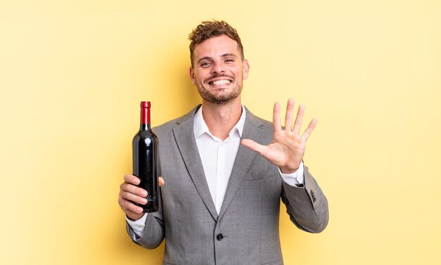 Hombre guapo joven sonriendo y mirando amigable, mostrando el número cinco. botella de vino concepto