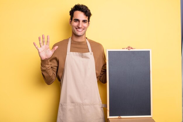 Hombre guapo joven sonriendo y mirando amigable, mostrando el número cinco. concepto de chef y pizarra