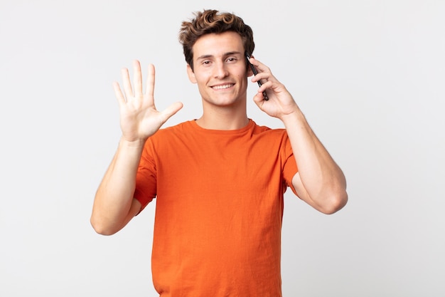 Hombre guapo joven sonriendo y mirando amigable, mostrando el número cinco y hablando con un teléfono inteligente