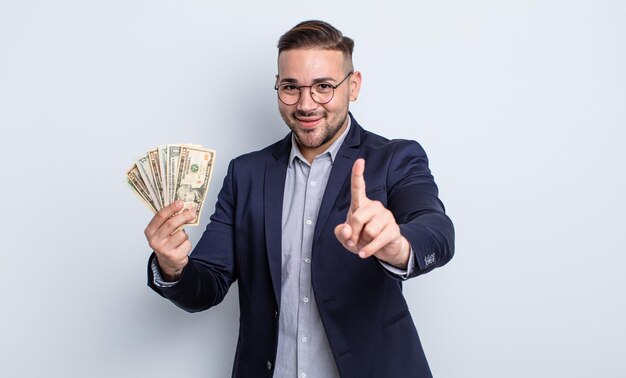 Hombre guapo joven sonriendo y mirando amigable, mostrando el número uno. concepto de billetes de dólar