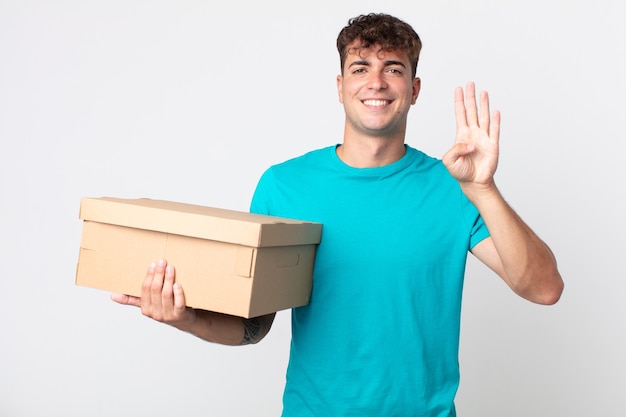 Hombre guapo joven sonriendo y mirando amigable, mostrando el número cuatro y sosteniendo una caja de cartón