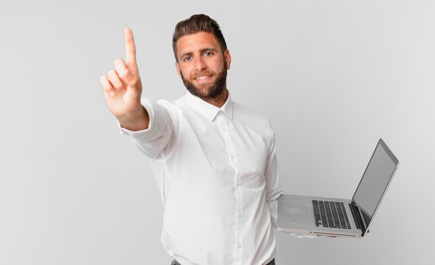 Hombre guapo joven sonriendo y mirando amigable, mostrando el número uno y sosteniendo una computadora portátil