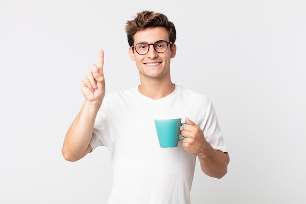 Hombre guapo joven sonriendo y mirando amigable, mostrando el número uno y sosteniendo una taza de café