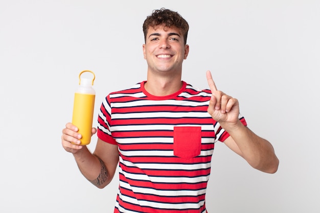 Hombre guapo joven sonriendo y mirando amigable, mostrando el número uno y sosteniendo un termo de café