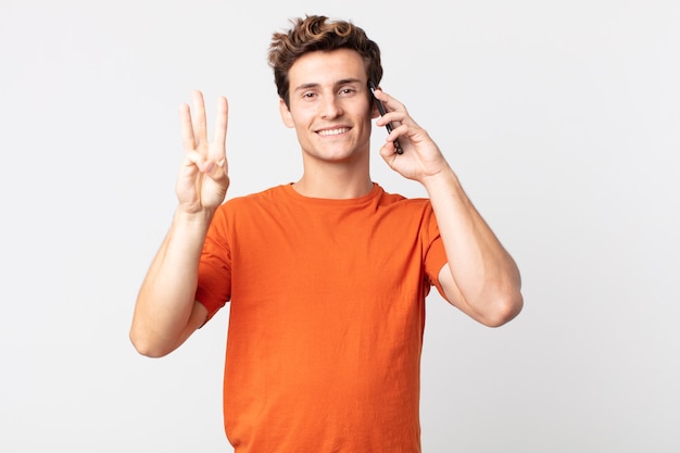 Hombre guapo joven sonriendo y mirando amigable, mostrando el número tres y hablando con un teléfono inteligente