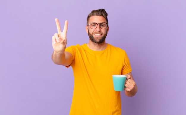 Hombre guapo joven sonriendo y mirando feliz, gesticulando victoria o paz. y sosteniendo una taza de café