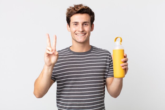 Hombre guapo joven sonriendo y mirando feliz, gesticulando victoria o paz y sosteniendo un termo de café