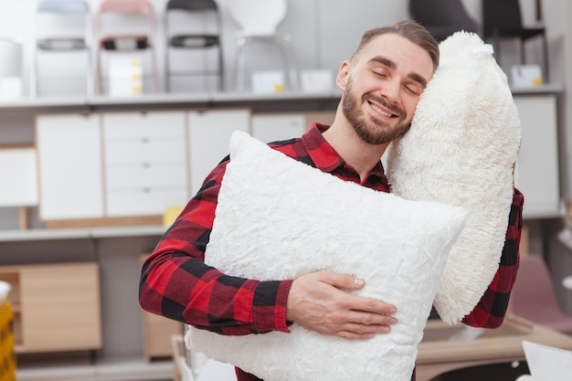 Hombre guapo joven sonriendo con los ojos cerrados, recostado la cabeza sobre una almohada esponjosa en la tienda de muebles