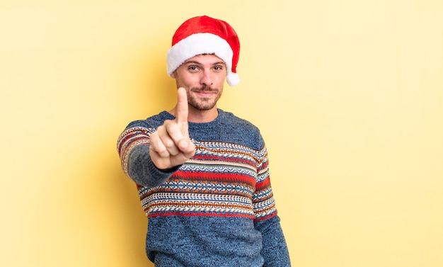 Hombre guapo joven sonriendo con orgullo y confianza haciendo el número uno. concepto de navidad