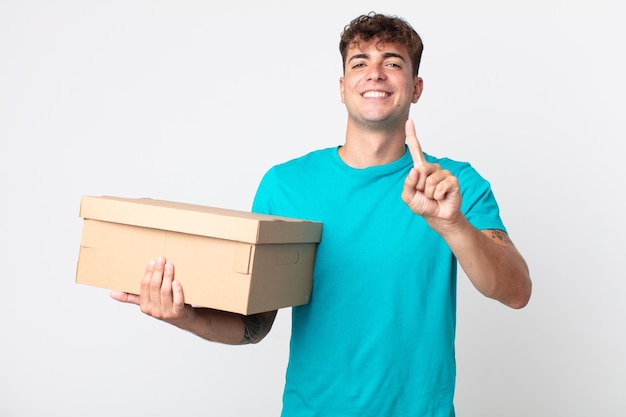 Hombre guapo joven sonriendo con orgullo y confianza haciendo el número uno y sosteniendo una caja de cartón