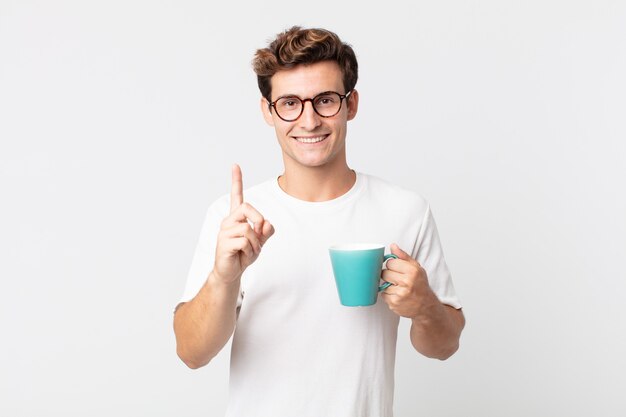 Hombre guapo joven sonriendo con orgullo y confianza haciendo el número uno y sosteniendo una taza de café