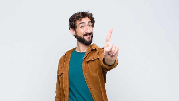 Hombre guapo joven sonriendo con orgullo y confianza haciendo la pose número uno triunfante, sintiéndose como un líder contra la pared plana