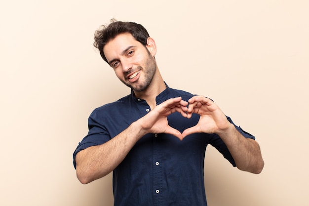 Hombre guapo joven sonriendo y sintiéndose feliz, lindo, romántico y enamorado, haciendo forma de corazón con ambas manos