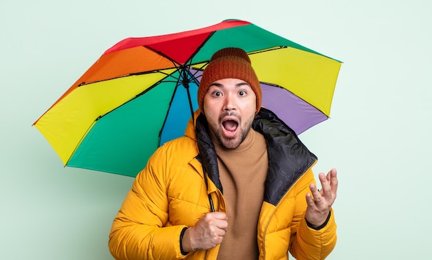 Hombre guapo joven sorprendido sorprendido y asombrado con un increíble concepto sorpresa de lluvia y paraguas