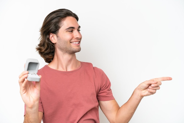 Hombre guapo joven sosteniendo un anillo de compromiso aislado sobre fondo blanco apuntando hacia un lado para presentar un producto