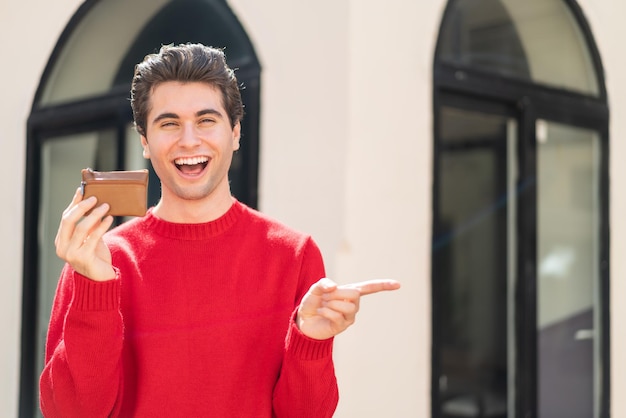 Hombre guapo joven sosteniendo una billetera al aire libre sorprendido y señalando con el dedo hacia un lado