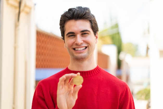 Foto hombre guapo joven sosteniendo un bitcoin al aire libre con expresión feliz