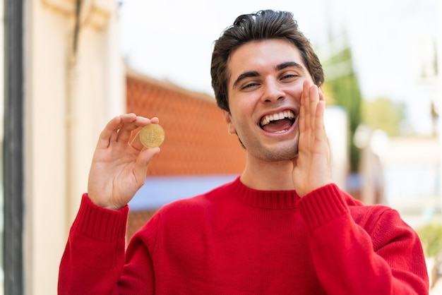 Hombre guapo joven sosteniendo un Bitcoin al aire libre gritando con la boca abierta