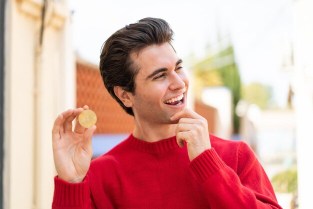 Hombre guapo joven sosteniendo un Bitcoin al aire libre pensando en una idea y mirando de lado