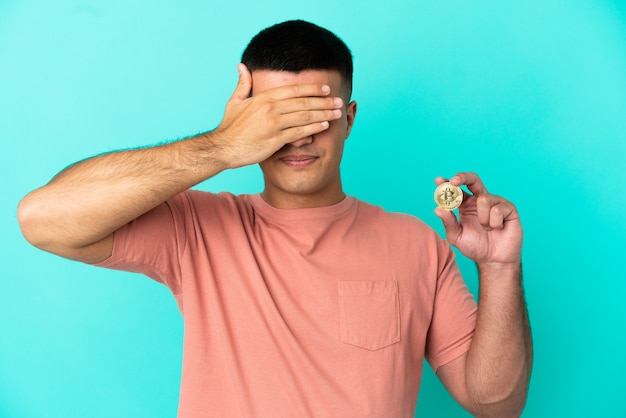 Hombre guapo joven sosteniendo un Bitcoin sobre fondo azul aislado cubriendo los ojos con las manos. No quiero ver algo