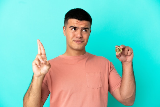 Hombre guapo joven sosteniendo un Bitcoin sobre fondo azul aislado con los dedos cruzando y deseando lo mejor