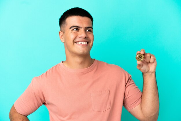 Hombre guapo joven sosteniendo un Bitcoin sobre fondo azul aislado posando con los brazos en la cadera y sonriendo