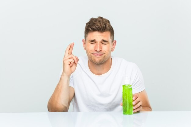 Hombre guapo joven sosteniendo una botella de aloe vera cruzando los dedos para tener suerte