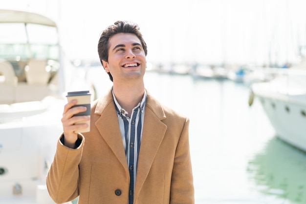 Hombre guapo joven sosteniendo un café para llevar al aire libre mirando hacia arriba mientras sonríe