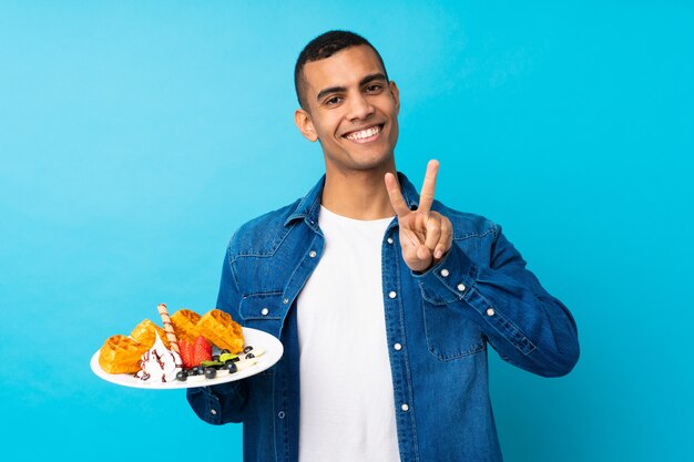 Hombre guapo joven sosteniendo gofres sobre pared azul aislado sonriendo y mostrando el signo de la victoria