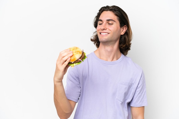 Hombre guapo joven sosteniendo una hamburguesa aislado sobre fondo blanco con expresión feliz