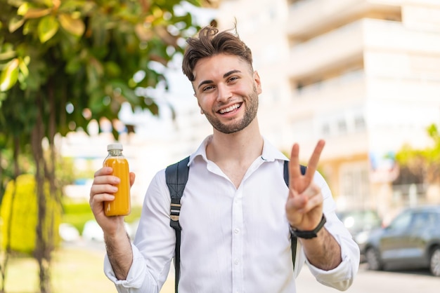 Hombre guapo joven sosteniendo un jugo de naranja al aire libre sonriendo y mostrando el signo de la victoria