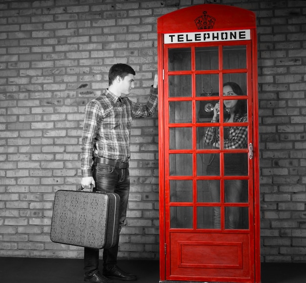 Hombre guapo joven sosteniendo la maleta llamando a la cabina telefónica con una mujer hablando dentro. Capturado con efecto de escala de grises.