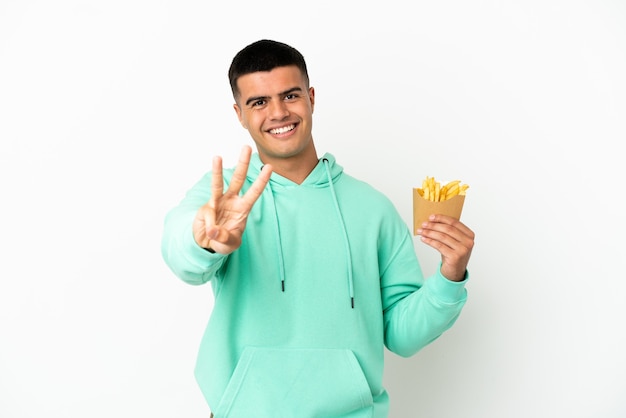 Hombre guapo joven sosteniendo patatas fritas sobre fondo blanco aislado feliz y contando tres con los dedos