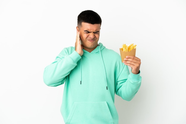 Hombre guapo joven sosteniendo patatas fritas sobre fondo blanco aislado frustrado y cubriendo las orejas